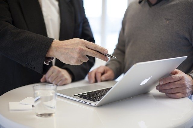 Zwei Menschen am Laptop, eine Hand weist auf das Macbook, im Vordergrund ein Glas Wasser.