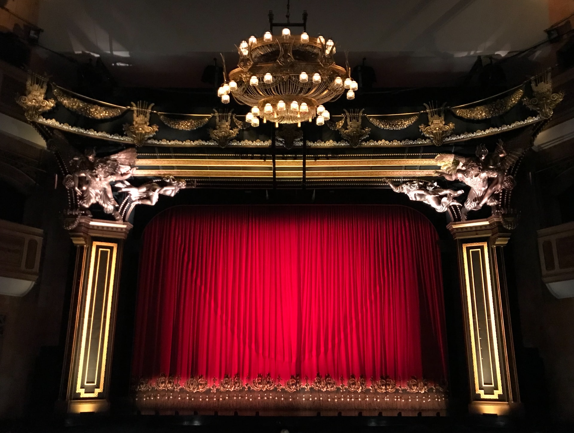 Sicht auf Theaterbühne mit rotem Vorhang. Oben prächtiger Kronleuchter, düstere Lichtstimmung.